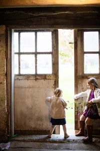 Two young girls, dressed in autumn attire, curiously peer out of an old rustic barn's window, surrounded by a vintage frame and a view of the outside world, in Metro Detroit.