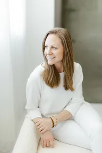 Light filled in studio professional headshot photo of a woman sitting down on a chair while she looks out of the window smiling.