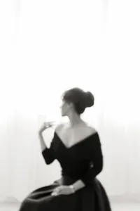 Beautiful black and white glamour shot of a mother dressed in a black gown that lays off the shoulders while she is looking out of the window as she sits in a photography studios in Dallas TX