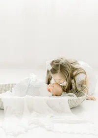Newborn baby boy laying in a basket swaddled on the floor as his big sister leans over him to kiss his forehead.