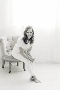 Black and white photo taken in a photography studio of a Dallas TX business woman sitting on a chair while she leans over to pose and smile at the photographer.