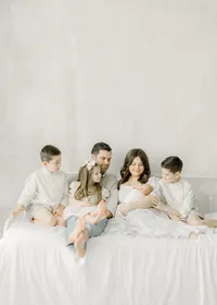 Beautiful Dallas TX family sitting on a bed together in a DFW photography studio while they are leaning in and as the siblings are looking at their newborn baby brother.