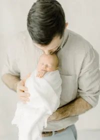 Father standing in a DFW photography studio holding his newborn baby boy as he leans down and kisses his head.