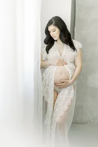 A pregnant mother dressed in a chiffon maternity gown while she is standing in front of a window looking down at her growing belly at a Dallas/Fort Worth photography studio.
