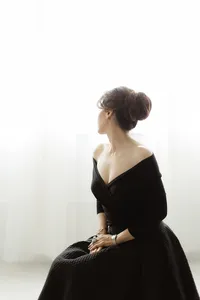 Beautiful glamour shot of a mother dressed in a black gown that lays off the shoulders while she is looking out of the window as she sits in a photography studios in Dallas TX