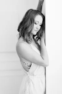 Light filled black and white photo of a woman posing for her boudoir session by leaning against a DFW studio wall while she looks down to the floor.