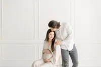 A maternity session taken at a Dallas/Fort Worth photography studio of an expecting mother sitting on a chair while her spouse is leaning over to kiss her forehead.