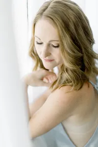 Close up photo of a woman posing by a window with sheer drapes with her hand elegantly places by her chest as she looks down over her shoulder for her boudoir session.