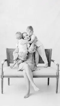 Black and white in studio photo of a mother sitting on a chair with her two boys snuggling her.