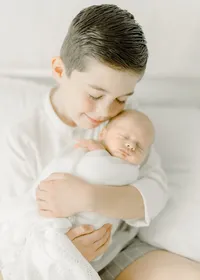 Big brother holding his newborn baby brother close as he smiles while sitting on a bed in a Dallas photography studio.