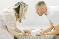 An in studio newborn portrait of a baby in a basket as her parents are holding her.