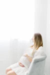 Maternity photo taken in a Dallas photography studio of a pregnant mom sitting on a chair looking outside of the window while she is holding her belly.