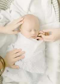 Newborn baby boy wrapped in a swaddle and tied with a light blue bow as his bigger siblings hold his hand and touch his head as he lays sleeping in a basket for his newborn session.