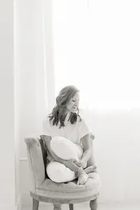 Black and white in studio professional headshot photo of a woman sitting down on a chair while she looks over her shoulder smiling.