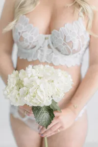Light filled boudoir photo of a woman wearing light blue lace lingerie set while she holds a bunch of fress ivory flowers in front of her.