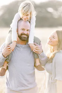 Close up family photo of a mother looking at her son as he is sitting on his dads shoulders.
