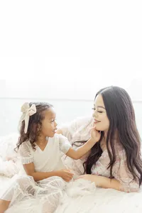 A light filled motherhood photo in a photography studio of a mother laying on a bed as her daughter gently holds her face as they smile at each other.
