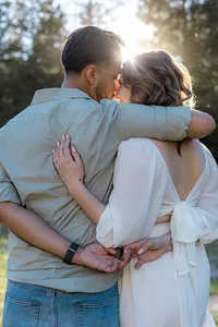 The couple embraces closely with the sun shining behind them, highlighting their affection as they hold each other with intertwined fingers.