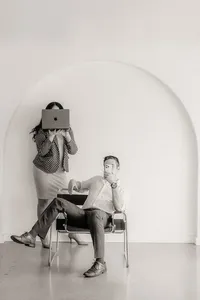 A man holds a phone in a chair while a woman stands behind on a laptop