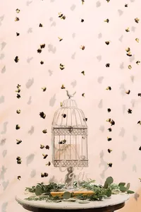 A birdcage cardholder labeled "Cards and Gifts" sits on a round table, adorned with greenery and surrounded by a backdrop of small dried flowers scattered across a light pink background.