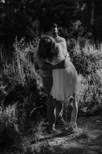 A black-and-white shot of the man lifting the woman in an affectionate embrace in a forest clearing.