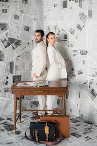 A couple stands back to back in front of newspapers