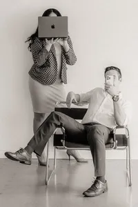 A man holds a phone in a chair while a woman stands behind on a laptop