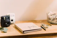 A wooden table setup featuring a guestbook labeled "guests" with two instant cameras, pens, and a glass bowl of snacks or mementos for guests.