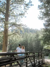 A couple leans on a rustic wooden bridge, smiling at each other, with tall trees and rolling hills as the backdrop, creating a peaceful, intimate scene in nature.