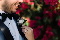 Close-up of the groom's boutonniere, a small white rose with greenery, as the bride’s hand gently adjusts it, with her engagement ring visible.