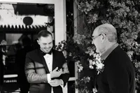 A candid black-and-white photo of the groom and his father sharing a laugh outdoors, with floral decor in the background.