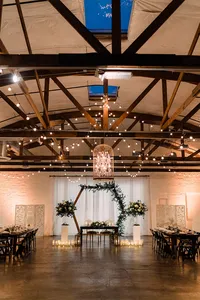 The wedding venue features wooden beams, string lights, and a chandelier hanging above the sweetheart table framed by greenery and candles, creating a romantic and rustic ambiance.