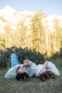 The couple lies side by side in the grass, holding hands and looking relaxed, surrounded by greenery and distant mountain views.