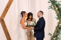 The bride and groom stand under a wooden arch decorated with greenery and white flowers, smiling at each other while the officiant behind them shares in their joyful moment.