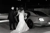 The bride, holding her bouquet, is escorted to a luxury car at night as she prepares to depart, with the groom holding the door open in a classic wedding send-off scene.
