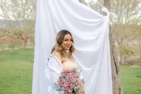 A pregnant woman stands in front of a sheet outdoors while holding flowers