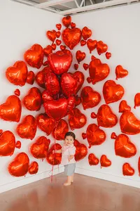 A baby boy stands in front of red heart balloons.