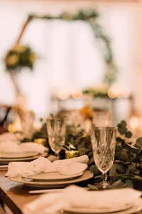 A close-up of an elegantly decorated wedding reception table with folded napkins, crystal glasses, and greenery centerpieces.