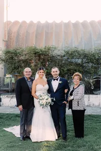 The couple stands outside with the groom’s parents, posing together on a green lawn with smiles, the bride holding a white floral bouquet.