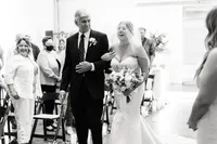 The father of the bride, smiling proudly, walks her down the aisle as she beams, holding a bouquet of white flowers and greenery, with guests looking on.