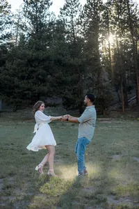 The couple holds hands and twirls playfully in a grassy clearing, with trees surrounding them.