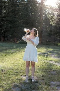A woman in a flowy white dress drinks from a clear bottle, standing in a sunlit field with sparkles of dust catching the light around her.