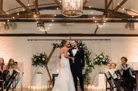 The bride and groom share a dance at their wedding reception, smiling and holding each other amidst an elegantly decorated venue with string lights and floral arrangements, while guests watch with smiles.