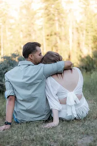 The man embraces the woman while they sit on the grass, her back to the camera; he places a gentle kiss on her head.