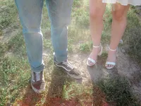 Close-up of a couple’s feet standing on grass, with the man wearing sneakers and jeans, and the woman in white high-heeled sandals and a light dress, emphasizing their contrasting casual and formal footwear.