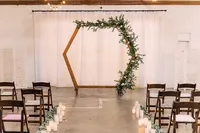 A minimalist hexagonal wooden arch decorated with greenery, placed in front of a white backdrop, with rows of chairs and a path lined with candles leading to it.