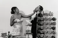 The bride playfully smears cake on the groom’s face while he laughs, with a wall of assorted donuts in the background