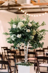 A lush floral arrangement of white roses, hydrangeas, and green foliage set in a tall glass vase, ready to adorn the ceremony space with chairs arranged in rows.