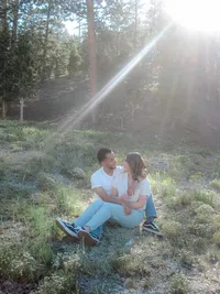 A couple sits in a grassy meadow with the sun casting a soft ray over them; they gaze affectionately at each other, creating a warm, romantic outdoor scene.