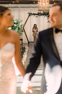 A blurred shot captures the groom watching his bride walk down the aisle, with candle-lit decor leading to a greenery-covered wooden arch in the background.